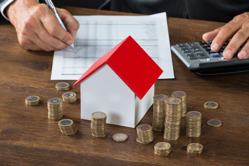 inflation vs home prices pile of coins in front of wooden house with person doing financial analysis with calculator
